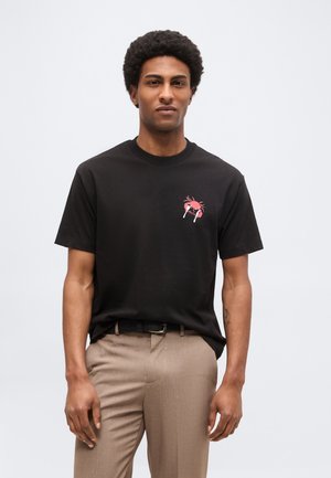 Hombre con cabello corto y rizado que lleva una camiseta negra con un pequeño gráfico de cangrejo rojo y pantalones beige, de pie frente a un fondo claro y simple.