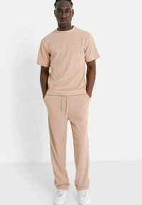Lichtbeige sweatsuit gemaakt van een gestructureerde stof, bestaande uit een t-shirt met ronde hals en joggingbroek met trekkoord, gecombineerd met witte sneakers.