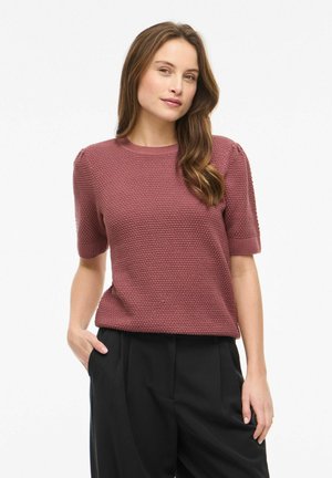 Kvinde med langt brunt hår iført en tekstureret, kortærmet mauve-sweater og sorte bukser med høj talje, poserer med den ene hånd i lommen.