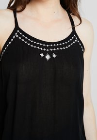 Top sans manches noir avec une finition texturée, ornée de motifs brodés blancs le long de l'encolure et d'une coupe décontractée.