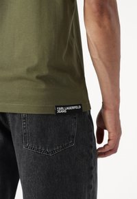 Olivno zelena majica s ravnim krojem, uparena s crnim trapericama. Na dnu lijevo se nalazi oznaka "KARL LAGERFELD JEANS".