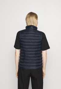 Granatowa kamizelka puchowa z wysokim kołnierzem, z pikowanymi przeszyciami i dopasowanym krojem. W połączeniu z czarną koszulką z krótkim rękawem i ciemnymi spodniami.