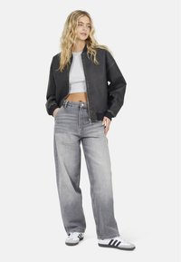 Giacca bomber nera con chiusura a zip e top grigio corto, abbinati a jeans grigi chiari a vita alta e gamba ampia, e sneakers bianche.