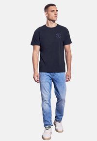 Marinblå bomull t-shirt med kort ärm, med en liten ljusblå grafisk design på bröstet. I kombination med ljusblå jeans och vita sneakers.