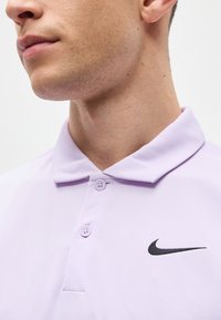 Κοντινό πλάνο ενός άνδρα που φοράει ανοιχτό μωβ πόλο Nike με μαύρο λογότυπο Swoosh στο στήθος και κουμπωτό γιακά.