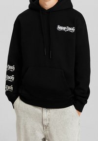 Felpa nera con testo bianco "Snoop Dogg" sul petto e ripetuto sulla manica sinistra, indossata con jeans grigi chiari e lavati.