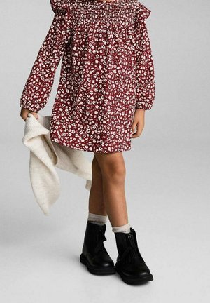 Robe florale rouge avec manches à volants, ornée de fleurs blanches sur un fond riche. Bottines noires à fermeture éclair et pull blanc en maille.