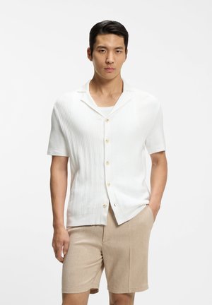 Mann trägt kurzärmeliges weißes Hemd und beige Shorts, steht mit einer Hand in der Tasche vor einem schlichten weißen Hintergrund.