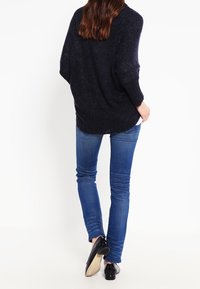 Námořnicky modrý převeliký pletený svetr s netopýřími rukávy, texturovanou látkou a žebrovaným lemem, kombinovaný s úzkými modrými džínami a černými botami.