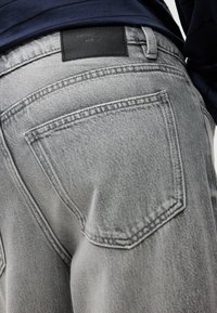 Artimos planas žmogaus, dėvinčio šviesiai pilkus džinsus, kuriuose matoma galinė kišenė ir juodas prekės ženklo lipdukas ant juostos.