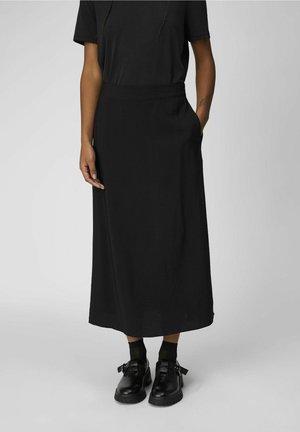 Jupe noire longueur cheville avec texture lisse, taille élastique et poches latérales, assortie à des chaussures et chaussettes noires.