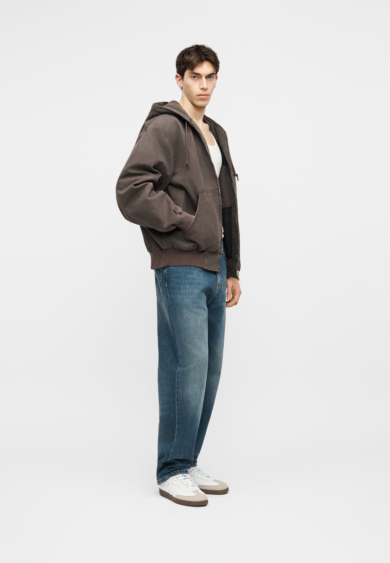 Giovane uomo con felpa grigia con zip, canottiera bianca, jeans blu e scarpe da ginnastica bianche, in piedi davanti a uno sfondo chiaro uniforme.