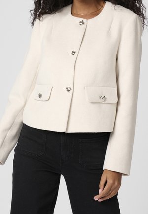 Kobieta ubrana w kremową wełnianą kurtkę z srebrnymi guzikami w kształcie serca i dwoma przednimi kieszeniami z klapkami, nosząca czarne spodnie.