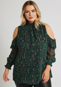 Grön och svart mönstrad blus med öppna axlar, smockad hals och manschetter. Tillverkad av genomskinligt tyg med en flödande design och avslappnad passform.