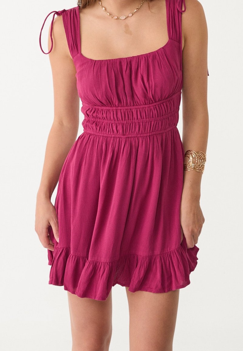 Femme portant une robe magenta sans manches, froncée, avec des bretelles nouées aux épaules, un ourlet à volants, un bracelet en or et un collier, sur un fond blanc.