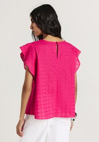 Top fucsia con motivi a diamante ricamati, maniche a sbuffo, leggera apertura posteriore con bottone e vestibilità morbida. Texture stropicciata visibile.