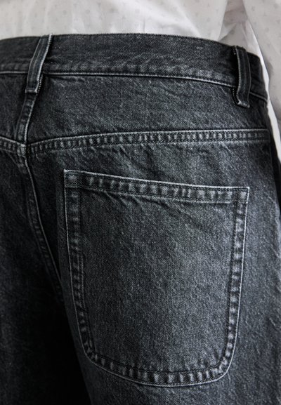 Svarta jeans med en bakficka, synlig sömnad och en texturerad tygkvalitet. Midjan har bälteshällor utan utsmyckningar.