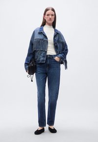 Τζιν μπουφάν με σχέδιο κρόσσια, λευκό ριμπ πλεκτό πουλόβερ, μπλε τζιν παντελόνι, μαύρα φλατ παπούτσια και μαύρη τσάντα με κορδόνι. Το μοντέλο στέκεται μπροστά σε ένα απλό φόντο.