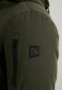 Primer plano de la manga de una chaqueta verde oliva con un bolsillo vertical de cremallera negra y un parche negro con un diseño naranja bordado.