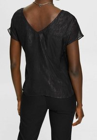 Černý krátký rukáv top s výstřihem do V, s texturovaným povrchem a jemnými metalickými vlákny. Nosit s černými kalhotami, které mají volný střih.
