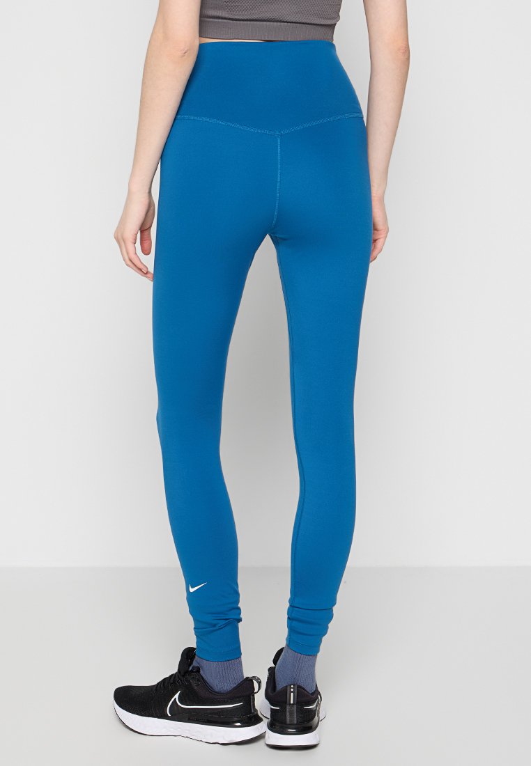 Person trägt blaue High-Waist-Leggings und schwarze Nike-Sneaker, steht mit dem Rücken zur Kamera vor einem weißen Hintergrund.
