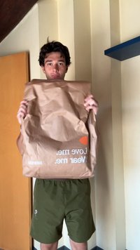 Saco de papel castanho com uma textura enrugada, apresentando texto branco e um gráfico de coração laranja. Segurado por uma pessoa em calções verdes.
