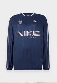 Mornariće plava dugom rukavu Nike majica s suptilnim prugama, sa velikim bijelim logotipom, tekstom "OD 1972" i izvezenim grbom na lijevoj strani prša.