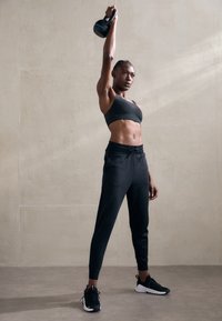 Svart atletisk sports BH og tettsittende joggebukser, kombinert med svarte løpetype sko. Holder en kettlebell over hodet i en treningspose.