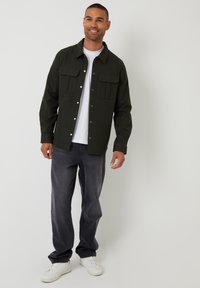 Donkergroene button-up shirt met twee borstzakken, gedragen over een wit T-shirt, gecombineerd met grijze jeans en witte sneakers.