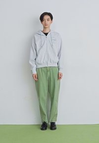 Noor naine seisab tasasel seinal, seljas heledashall tõmblukkidehoodie, laiad rohelised püksid ja mustad kingad, vaadates otse ette.