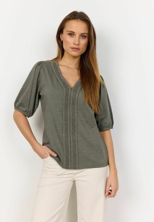 Olive green blouse with a V-neck and lace detailing down the front. It features puffed sleeves and a relaxed fit, paired with cream trousers.