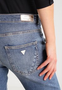 Ljusblå denimjeans med en slitagetekstur, som har en triangulär silverlogotyp på bakfickan och en metallisk varumärkesetikett på midjebandet.