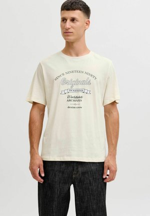 Lys beige crew neck t-shirt med sort trykt tekst, der læser "Siden Nitten Halvfems Originals Jack & Jones Verdensomspændende Arkiver." Korte ærmer.