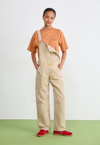Salopette en denim beige avec une seule bretelle, associée à un t-shirt orange. Accessoires : baskets rouges et coutures plates simples.
