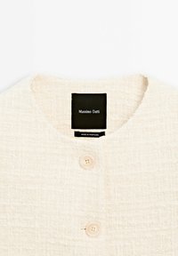 Veste à texture crème avec un col rond, deux boutons crème et une étiquette noire portant la marque Massimo Dutti, fabriquée au Portugal.