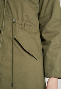Veste vert olive avec une poche poitrine proéminente, une taille à cordon de serrage et un tissu texturé. Le matériel argenté met en valeur le design.