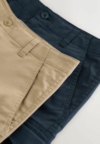 Gros plan sur des pantalons beige et bleu marine foncé pliés, montrant les ceintures, boutons, passants de ceinture et poches avant sur une surface blanche.