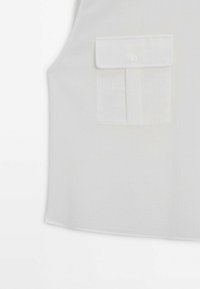 Haut blanc sans manches avec une seule poche poitrine dotée d’un rabat et d’une fermeture par bouton du côté droit.