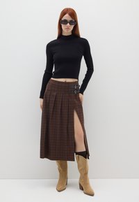 Top court noir à côtes, jupe à carreaux marron avec fente latérale et détails de boucle, associée à des bottes à talons en suede beige, sur fond blanc.