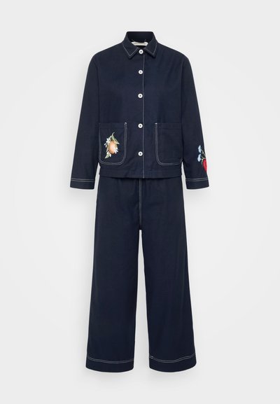 Klasisks tumši zils kokvilnas divdaļīgs kostīms, kas sastāv no uz pogām aizverama žaketes ar divām kabatām un ziedu izšuvumiem, apvienots ar platām biksēm.