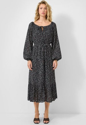 Femme blonde portant une robe midi noire à motif floral avec des manches longues bouffantes, une taille cintrée et des escarpins noirs à bout ouvert, debout contre un mur blanc uni.