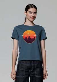 T-shirt en coton turquoise, ornée d'un graphique de coucher de soleil circulaire en orange et rouge avec des silhouettes d'arbres en bas. Manches courtes.