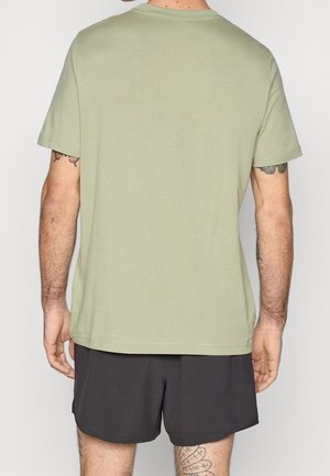 Mężczyzna ubrany w jasnobrze zielony t-shirt z krótkim rękawem i czarne krótkie spodenki, stoi tyłem do kamery, pokazując tatuaże na ramieniu i nodze.