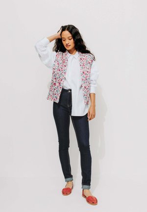 Gilet floral sans manches sur une chemise blanche, associé à un jean skinny noir et des chaussures slip-on tissées rouges. Cheveux noirs et ondulés avec un fond neutre.