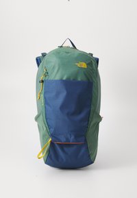 Mochila de nylon verde y azul con forma redondeada, que cuenta con un bolsillo frontal con cremallera, correas ajustables y un tirador de cremallera amarillo. Logo de The North Face visible.