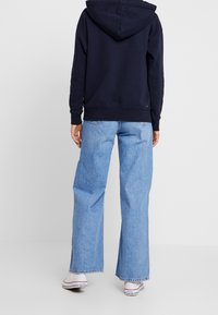 Sudadera con capucha en color navy, con bolsillo tipo canguro, combinada con jeans de pierna ancha en azul claro. Los jeans tienen un dobladillo deshilachado y costuras sutiles.