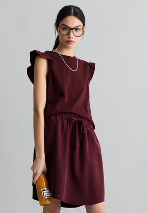 Jeune femme aux cheveux noirs attachés en queue de cheval, portant des lunettes écaille de tortue et une robe bordeaux, tenant une bouteille de boisson orange.