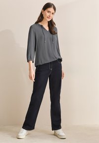 Blusa gris ligera con cuello en V y cordones, combinada con jeans de pierna recta de color azul oscuro y zapatillas blancas.