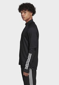Zwart langemouwen sportshirt met hoge kraag, gestructureerde stof en witte streepaccenten op de mouwen, gecombineerd met zwarte broek.