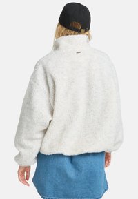 Bela, puhasta flis jakna z visokim ovratnikom in spuščeni rameni. Ima majhno oznako logotipa na hrbtni strani. Oblečena je čez denim krilo.